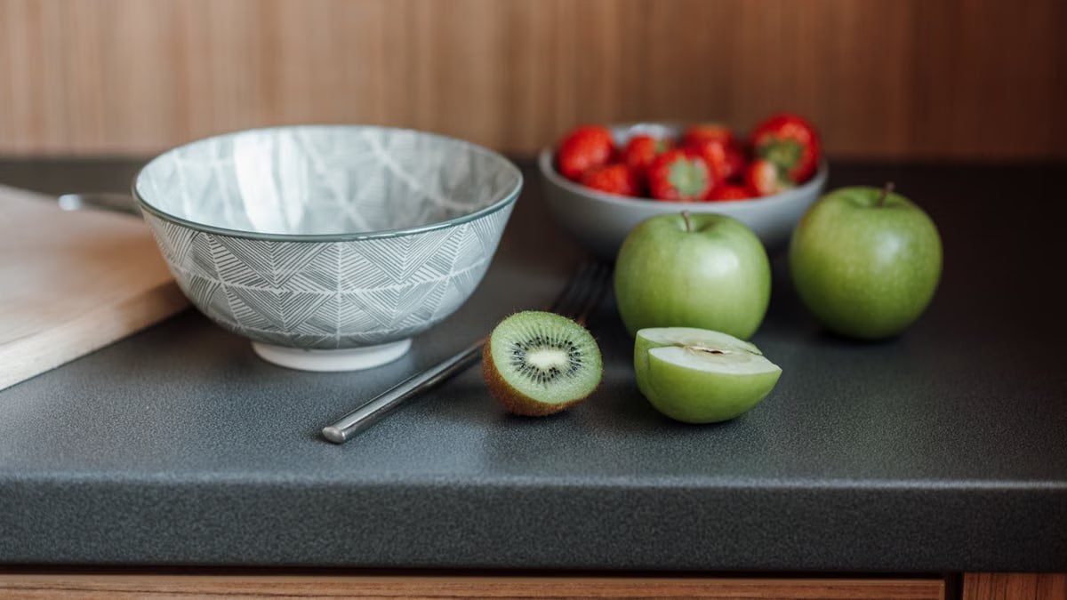 Table with bowl near fork and green apples with kiwi and cutting board near strawberries in dishware in light kitchen